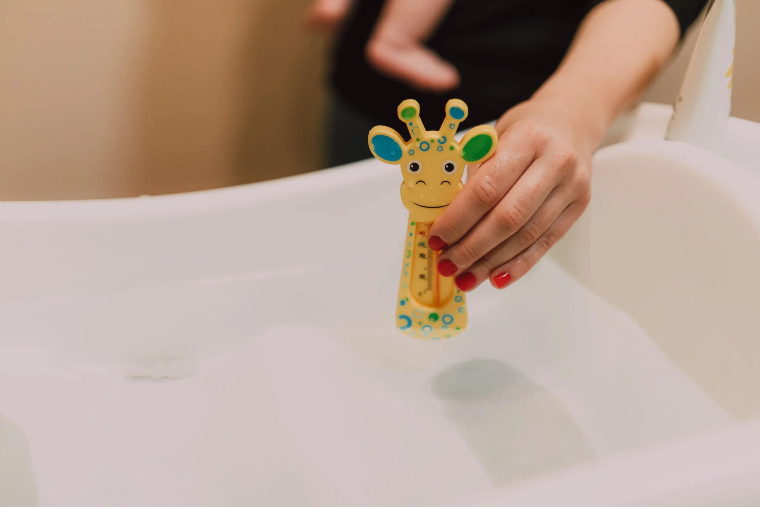 Une main de parent tenant un thermomètre jaune en forme de girafe au-dessus d'une baignoire pour bébé.