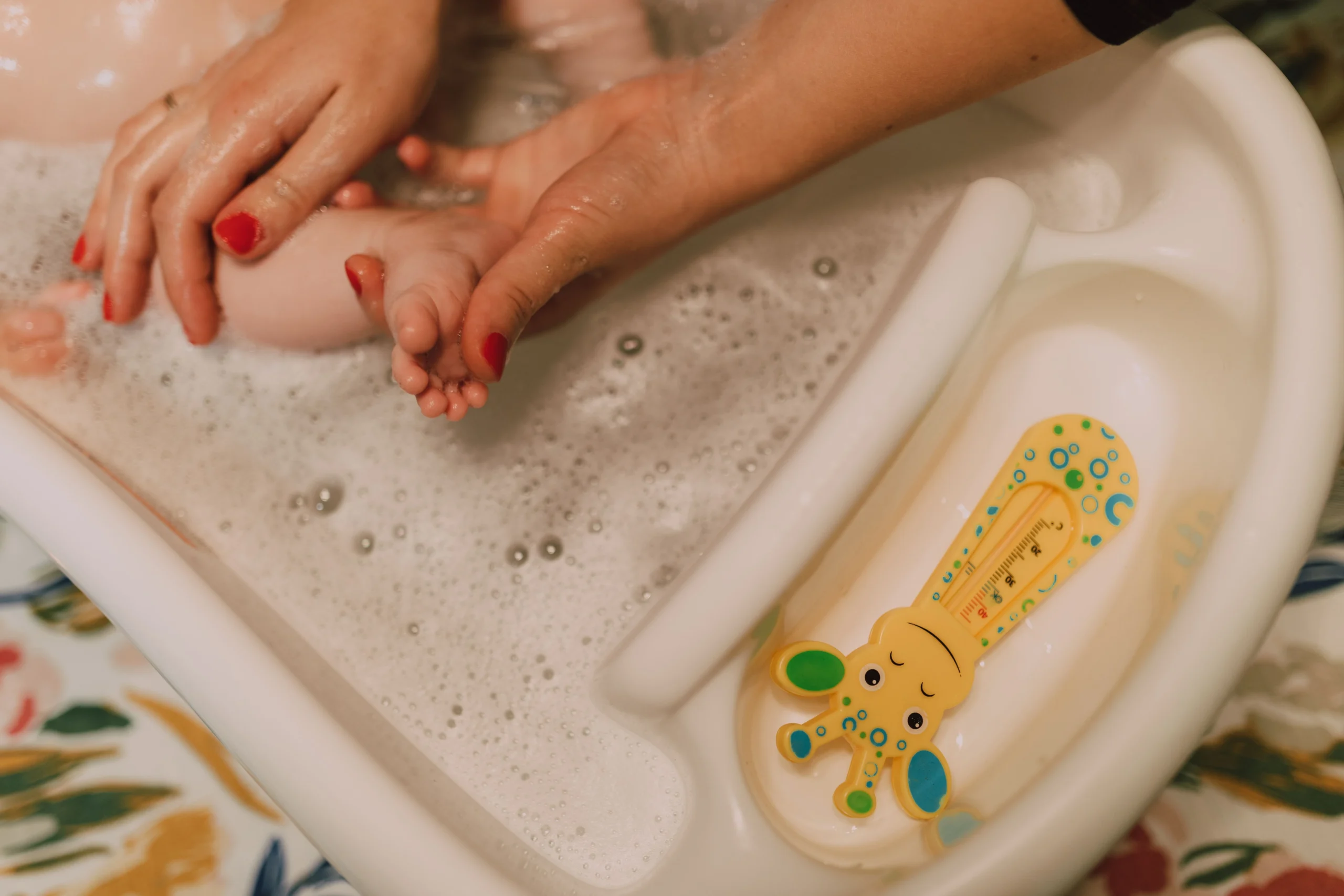Vue en plongée sur des mains lavant doucement un bébé avec le thermomètre girafe posé dans la baignoire.