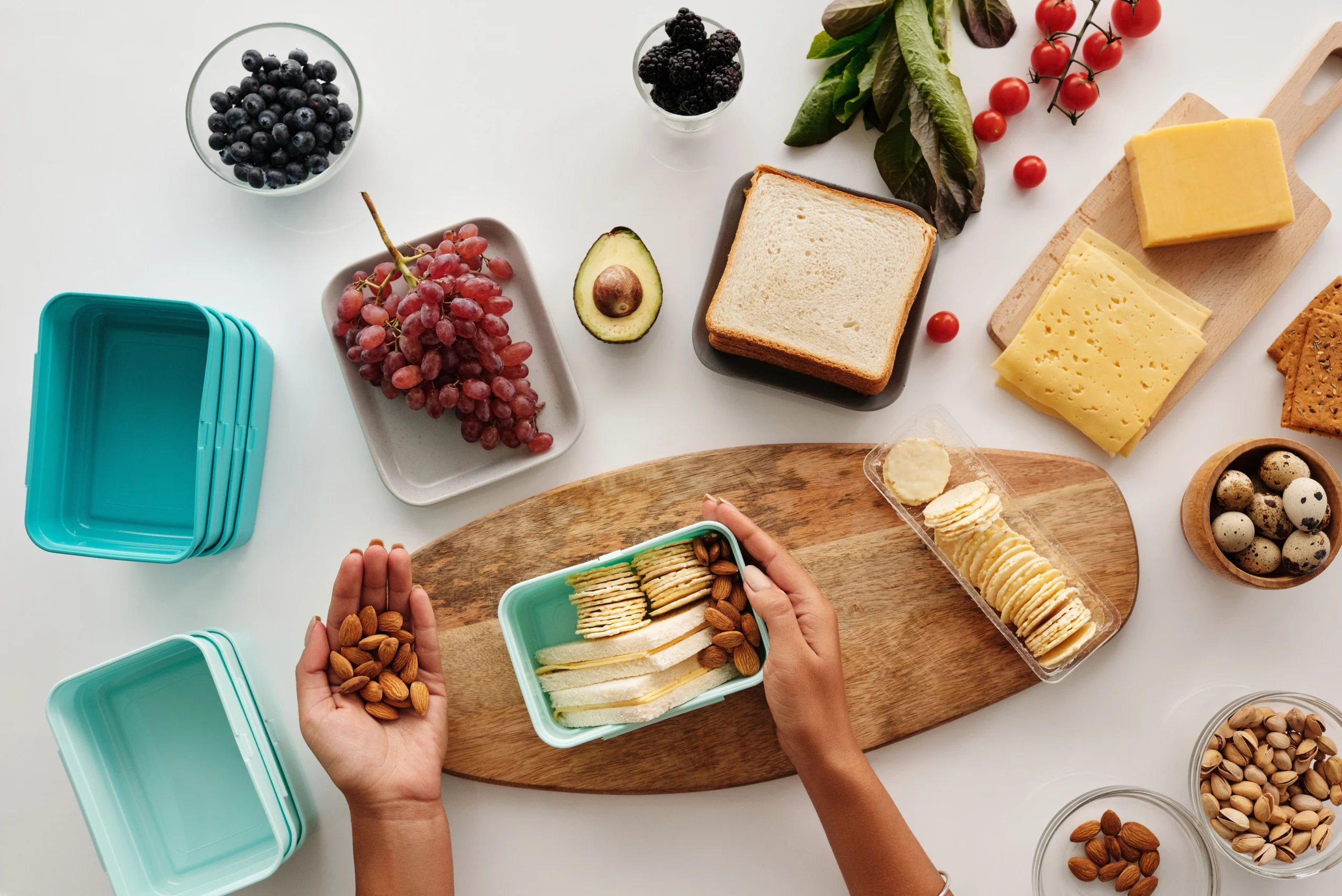 Des mains préparant une boîte repas avec des amandes, du pain de mie, du fromage et des fruits sur une table.